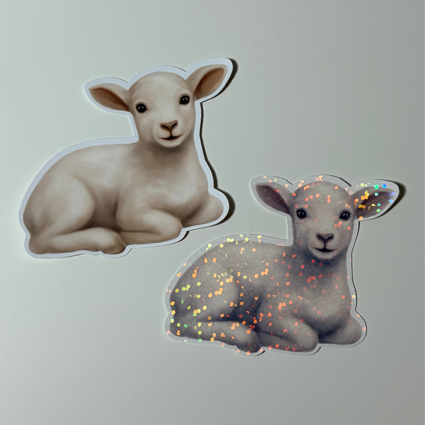 Two stickers of lambs, one plain and one with glitter, on a light gray background.